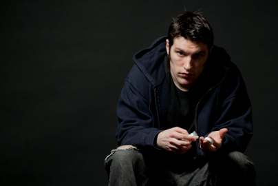 A white male with dark hair wearing a hoodie holds a pill bottle and stares toward the camera. 