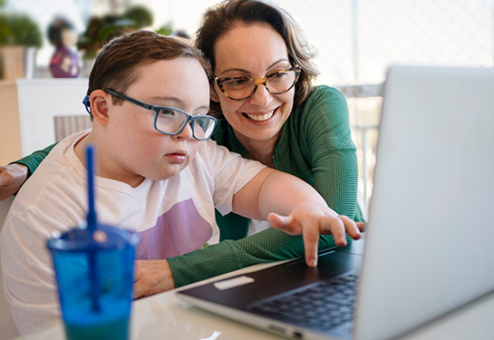 A mother and her son, who has Down syndrome, are sitting at a table working on a computer both wearing glasses