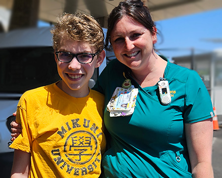 A blonde boy with a brown-haired woman wearing scrubs stand in front of the hospital entrance.