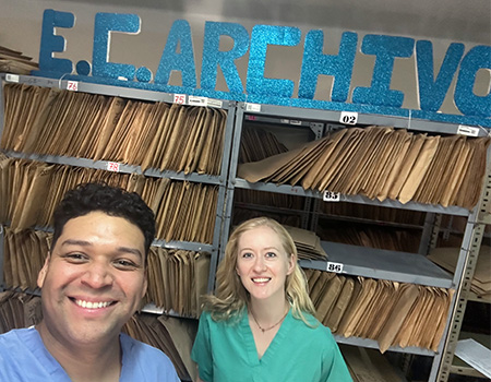 A male and a female neurological surgery resident in scrubs smile before shelves of patient records they began digitizing