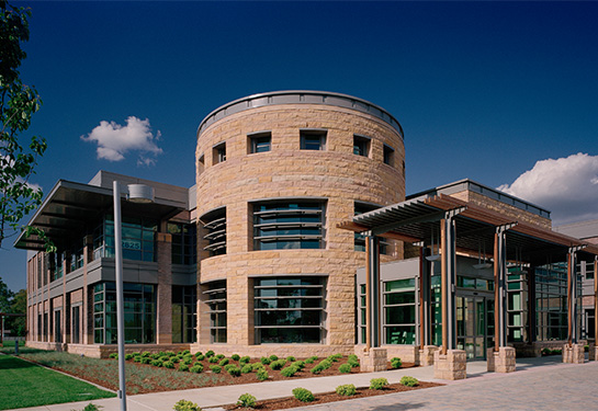 A large sandstone and glass building flanked by bushes and trees. 
