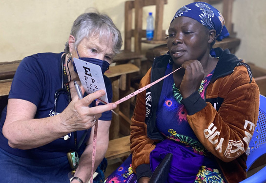 Nurse practitioner holds card with letters to test sight of Kenyan woman who holds a tape measure in her hand while reading off the card
