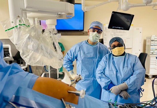 Two people stand side by side wearing blue scrubs, surgery caps, gloves and masks in the operating room.