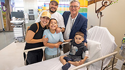 Young boy sits in hospital bed while physician and older brother lean over him and father and mother watch in the background.