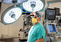 Male surgeon stands in center of operating room under large operating room lights, wearing a yellow surgical mask and a beige high-tech visor.